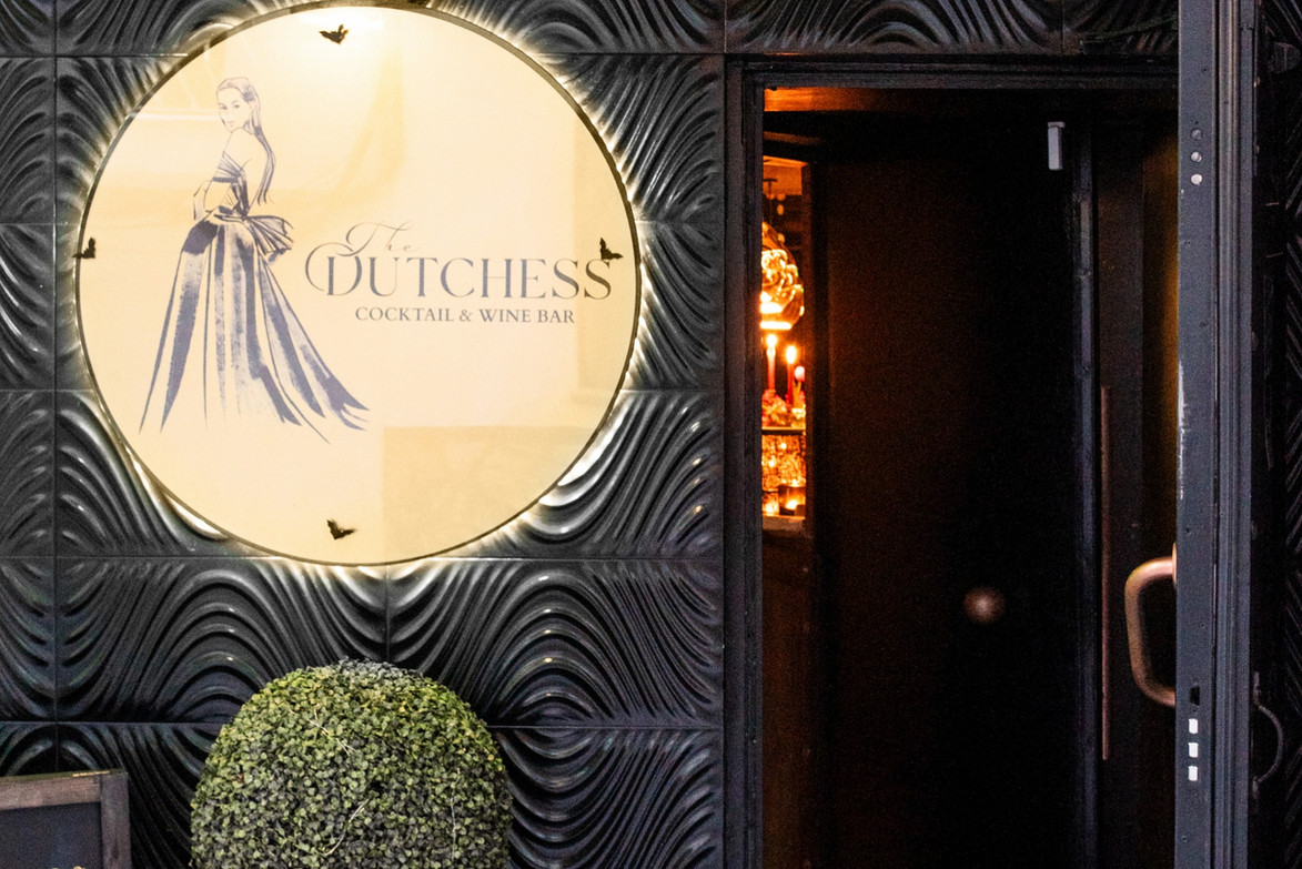 Entrance to The Dutchess bar featuring a black wooden door, round logo sign with The Dutchess figure and text, and a black pot with a green bux tree plant.