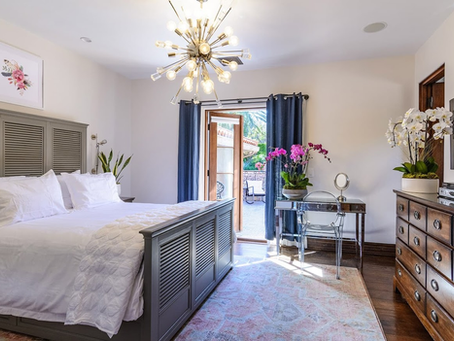 Elegant bedroom with modern chandelier, gray paneled bed, fresh white bedding, and vibrant orchid flowers near glass desk and dresser