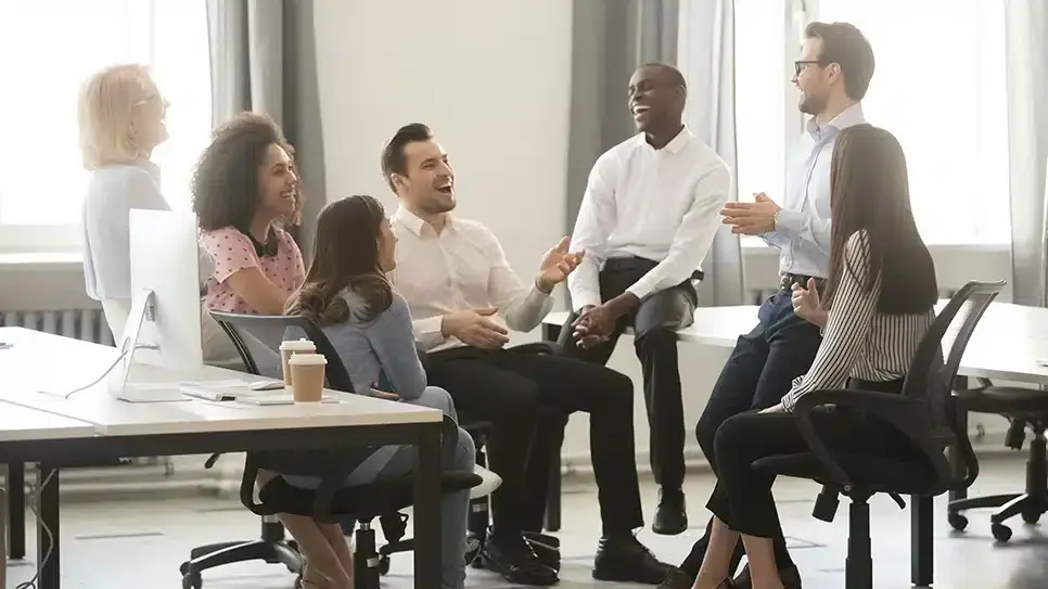 A diverse group of seven people sit and laugh in a bright office setting. They're engaged and appear to be having a lively, joyful discussion.
