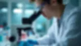 Researcher wearing goggles and gloves examines a sample through a microscope in a lab. Blue and white dominate the setting.