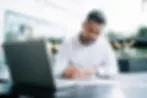 Man in white shirt writing at a black table with a laptop outdoors. Blurred city background, focused expression, modern ambiance.