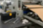 Industrial machine feeds corrugated cardboard in a factory, with a toolbox, wrenches, screwdriver, and gloves on a workbench.