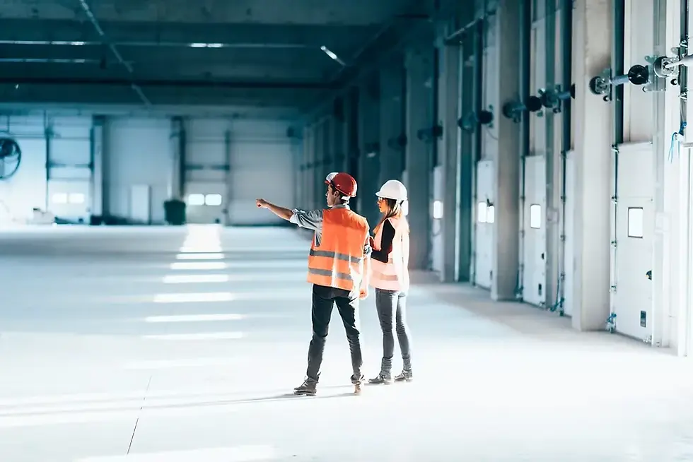 Two people in hard hats and vests stand in a large, empty warehouse. One points forward. The scene is brightly lit with blue-gray tones.
