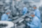 Workers in blue uniforms and hairnets operate machinery in a sterile factory, organizing white containers. Monitors display data.