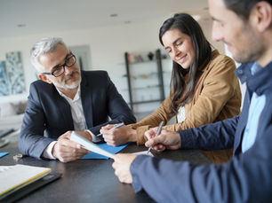 Three people review documents at a table in a bright office, smiling and writing notes during a friendly meeting.