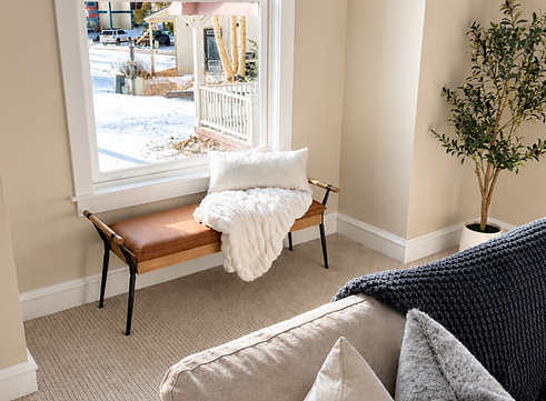 Staged window nook at Wellington with a leather and wood bench, a cream knit pillow and throw blanket, a tall faux olive tree, and a snowy street view through the window.