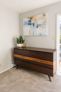 Staged bedroom corner at Deer Path with a dark walnut dresser featuring a lighter wood inlay stripe across the drawers, a white ceramic planter with a small plant and a gold knot decor piece on top, a blue, tan, and white abstract painting on the wall above, beige carpet, and a glass door leading outside.