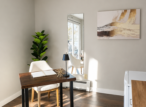 Staged home office corner at Deer Path with a light wood desk and matching wood-and-cream upholstered chair, an open book and small gold star-shaped decor on the desktop, a tall leaning mirror, a fiddle leaf fig plant, a black table lamp, a large gold and cream abstract painting on the wall, and dark wood flooring.
