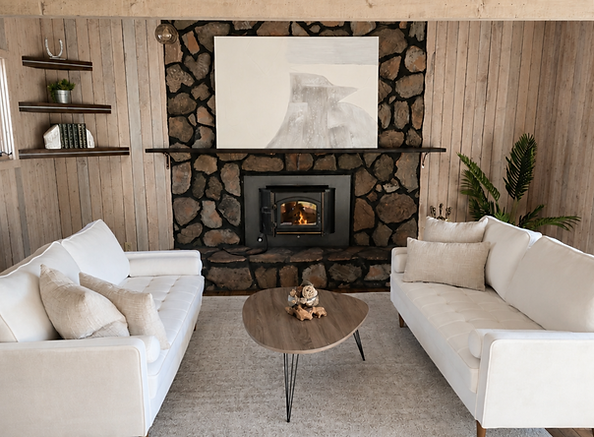 Staged living room at Deer Path with vertical wood-paneled walls, a stone fireplace with a black wood-burning insert, an abstract black and white painting above the mantel, dark wood floating shelves styled with books and a small plant, two white tufted sofas, an oval wood coffee table with a decorative sculpture, a neutral patterned area rug, and a leafy potted plant in the corner, by Fresh Air Styling Co.