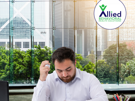 A man in a white shirt sits at a cluttered desk, surrounded by crumpled papers and looking overwhelmed — reflecting the challenges of focus and frustration often experienced with ADHD. Office buildings and trees are visible through a large glass window. The Allied Behavioral Health Solutions logo is shown in the top right corner.