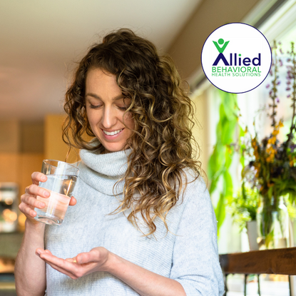 Woman holding a glass of water and looking at a supplement pill in her hand, considering taking a dietary supplement for wellness.