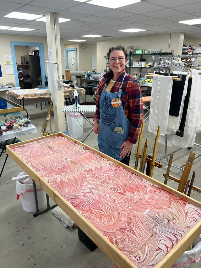 In a classroom setting, a woman stands near a water marbling tray that has two pieces of silk fabric laying on the surface of the solution.