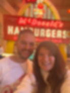 Two people smiling in front of a retro McDonald's sign with neon colors. The sign reads "Hamburgers" and "Over 1 Million Sold."