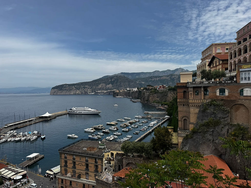 Coastal view with buildings, a marina of boats, and a large ferry named SNAV. Cliffs and mountains in the background under a partly cloudy sky.
