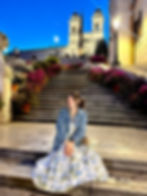 Woman in a floral dress and denim jacket sits on steps with flowers, smiling at dusk. Illuminated church in the background. Calm setting.