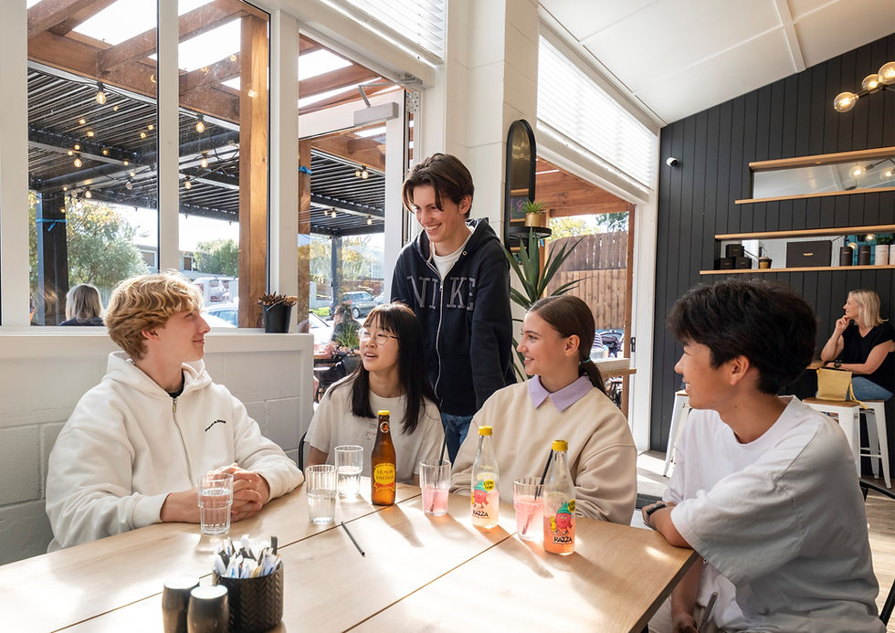 Five diverse young international students at Howick College chatting and laughing at a bright cafe table.