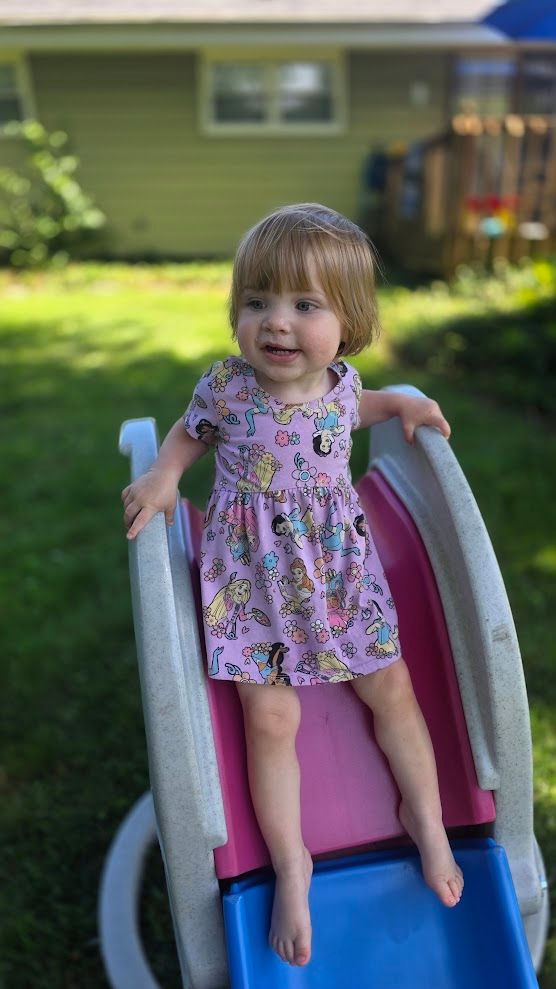 Toddler in a pink dress sitting on a backyard slide.