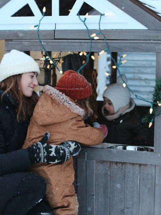a mother hugging her child in the snow near a backyard playhouse while another child plays inside the playhouse