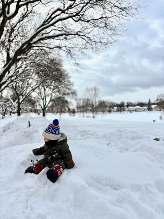 a baby wearing snow bunting, booties, and a hat, sitting on top of a huge pile of snow