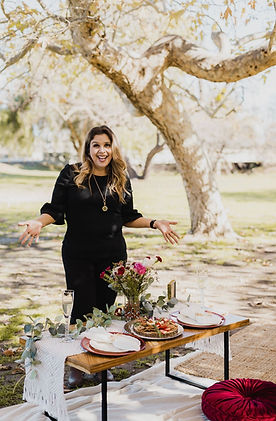 Woman in a black dress presenting a low picnic table set with a white woven runner, dark red chargers, white plates and napkins, clear glassware, greenery, pink and dark red flowers, floor cushions, and layered woven rugs.