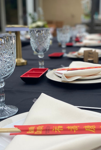 Dinner table setting with a black tablecloth, clear goblets, white plates and napkins, and chopsticks paired with a small sauce dish.