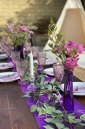Wooden table decorated with white plates, purple napkins, a purple runner, candlesticks, purple glass goblets, purple vases with flowers, and greenery.