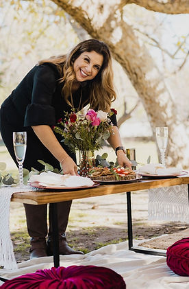 Woman in a black dress smiling at the camera while reaching for a plate of food at a low picnic table set with a white woven runner, dark red chargers, white plates and napkins, clear glassware, greenery, pink and dark red flowers, floor cushions, and layered woven rugs.