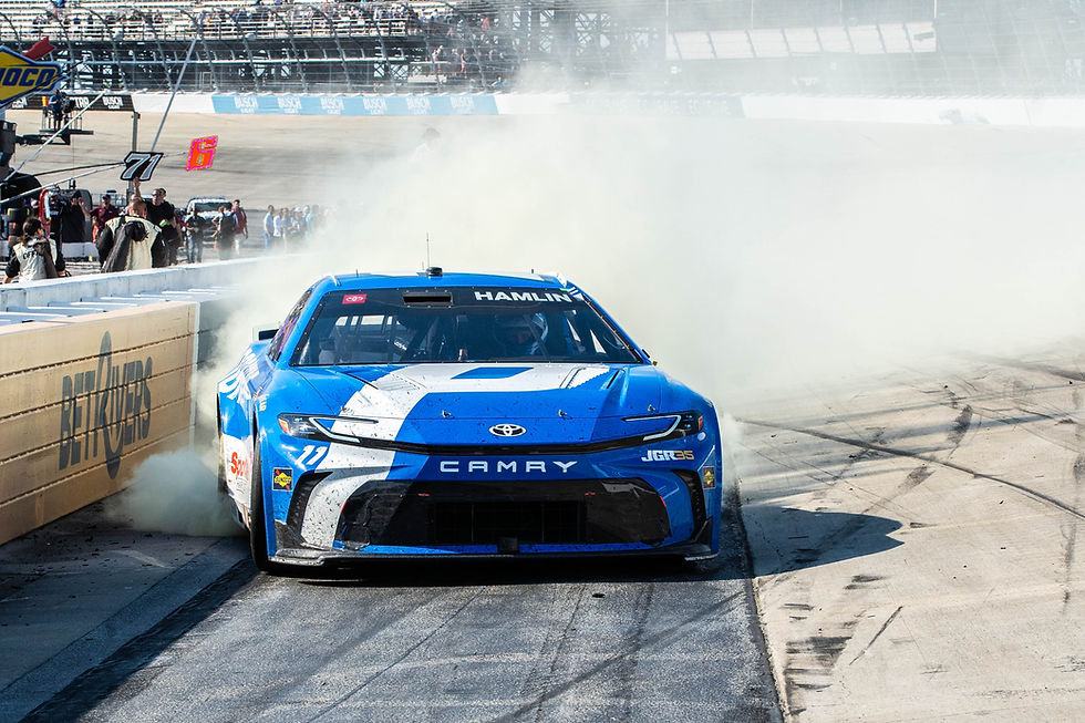 Blue race car performing a burnout, smoke billowing around. "Camry" and "Hamlin" visible on the hood. Crowd in the stands watches.