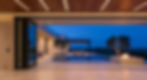 View through a massive pivot door of an Axiom Modern home at dusk, showing a seamless transition from the great room to a terrace with a negative-edge pool and sunken fire lounge.