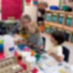 An adult wearing a teacher aide apron sits next to kids at a craft table. On the table are eggs and food coloring dye.