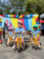 3 children on tricycles with rainbow banner in background.
