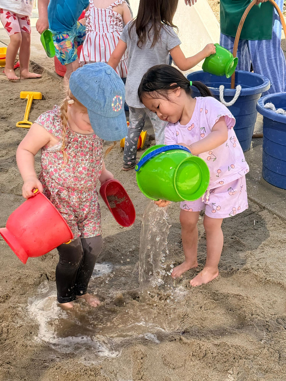 2 girls have fun dumping buckets of water on their feet in the sand.