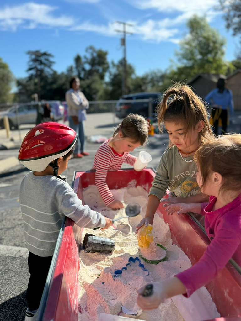 Kids gather around a red table filled with flour, cups, ladles, and sieves.