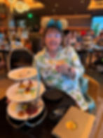 Woman in floral shirt with gold ears holds a drink at a restaurant. A three-tiered tray of desserts is on the table. Warm lighting.
