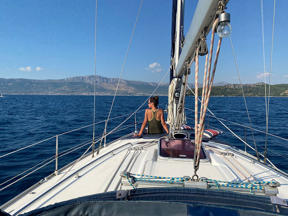 Woman sitting on a yacht deck facing the sea and mountains. Clear blue sky, calm waters. Peaceful and sunny atmosphere.
