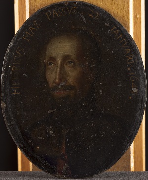 Oval portrait painting showing a man with a moustache and goatee staring to the left of viewer with inscription in yellow paint in an arch around his head, dark and inscription harder to read, and noticeable chips to the paint
