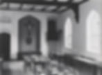 Black and white photo showing a room with set tables and a large crucifix at end of room middle of the photograph.