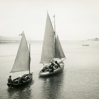 Black and white photo of two sail boats out on open water. Each contains several people in dark clothing.