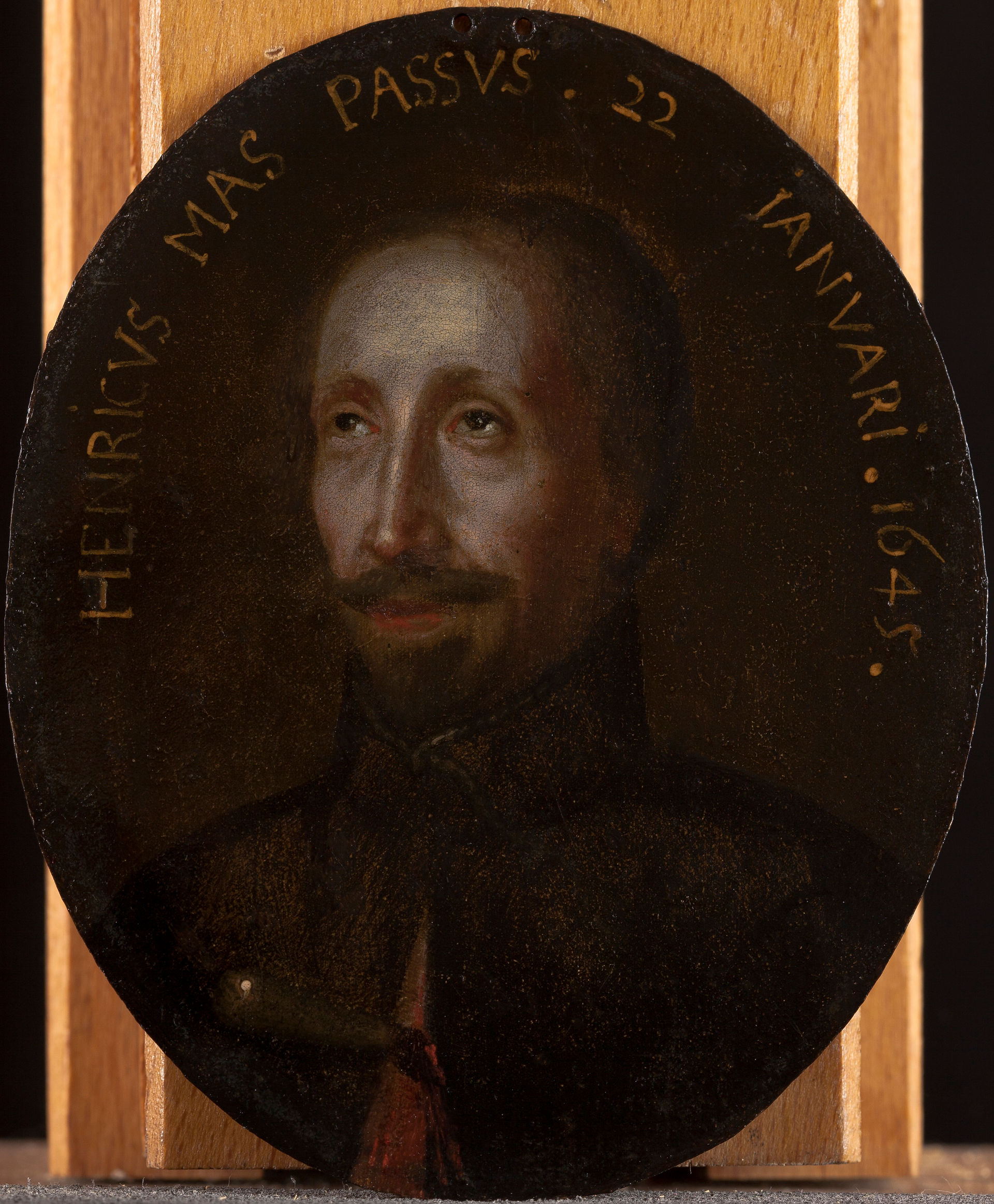 Oval portrait painting showing a man with a moustache and goatee staring to the left of viewer with inscription in yellow paint in an arch around his head, brighter and without any noticeable chips in paint