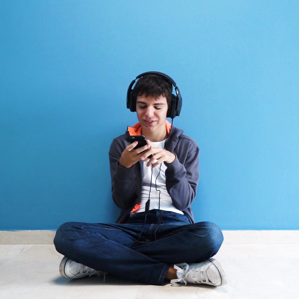 A young boy with headphones sits on the floor against a blue wall and plays on his phone