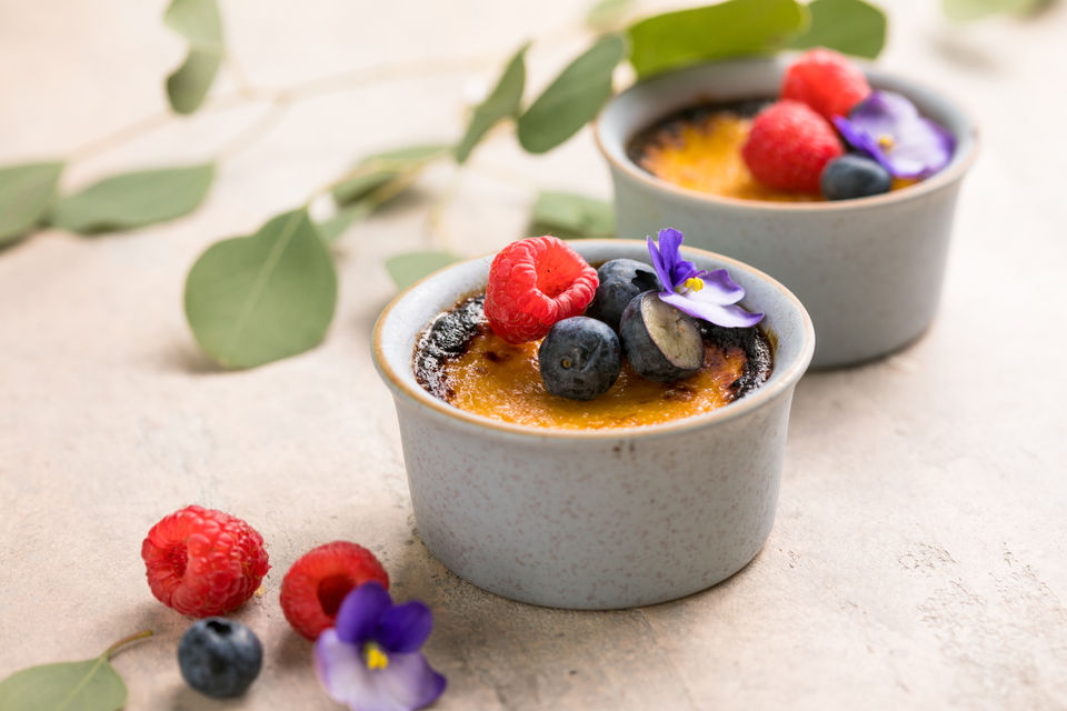 Dos crème brûlée con frambuesas, arándanos y flores comestibles sobre mesa.