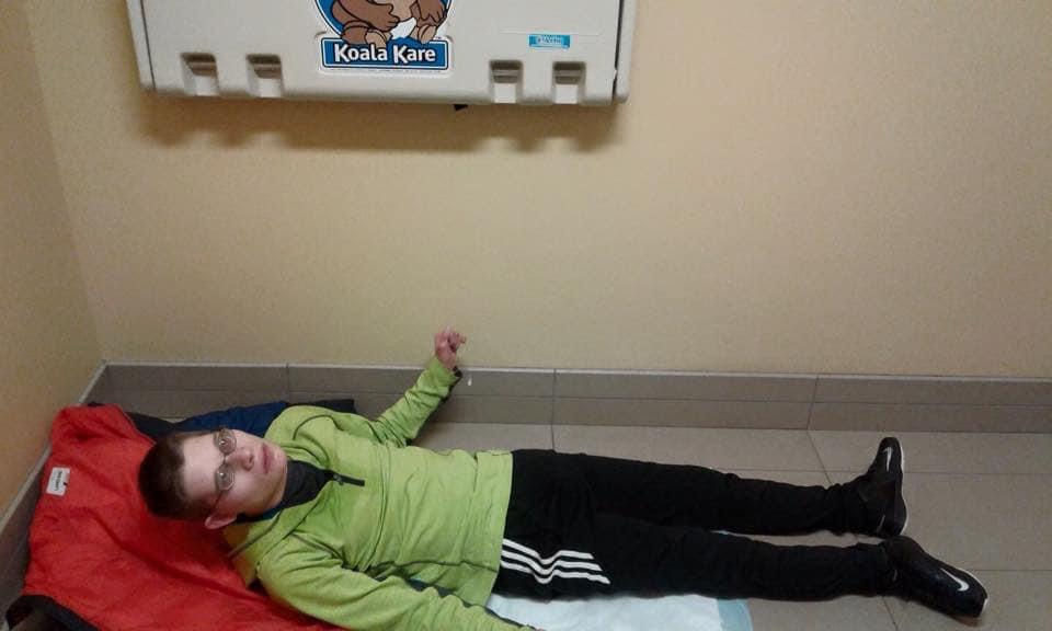 19 year old young man lies on the restroom floor underneath a baby changing table that he is too big for.