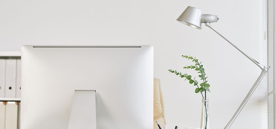 The back of a silver desktop screen. Behind it is a white shelf with white file folders. To the right is a clear vase with two stems of a small, leafy plant. A white bendy lamp is to the right, angled towards the computer.