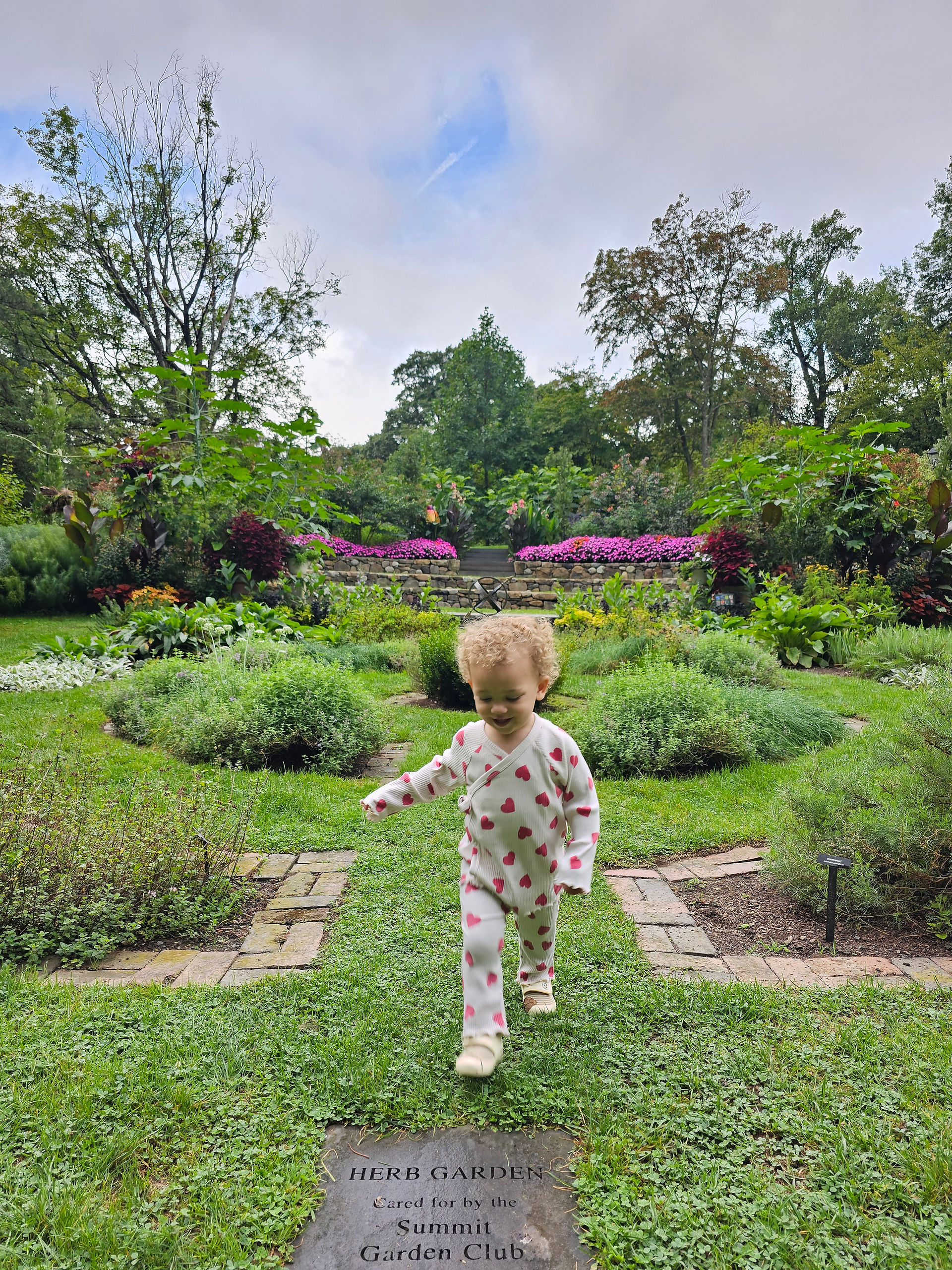 Rose, a toddler with curly hair, running happily across a grassy path in the center of the geometric Herb Garden at Reeves-Reed.