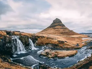 Et fantastisk landskap med et fossefall som renner ut i en elv, med utsikt til Kirkjufell-fjellet på Island. Den ruglete terrenget er dekket med gyllent gress, og en enslig figur kan sees i det fjerne. Himmelen er overskyet, noe som gir en dramatisk atmosfære til dette ikoniske naturlandskapet.