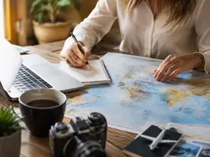 Traveler planning a trip at a bright desk workspace with a world map, laptop, notebook, camera, passport, and coffee cup, illustrating the process of using a travel planner versus booking independently.