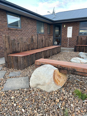 Close-up of large stone carved for seating with an aged hardwood timber beam, alongside kwila seating set on railway sleepers, built by Heartland Landscapes in Christchurch as part of a rustic fire pit area.