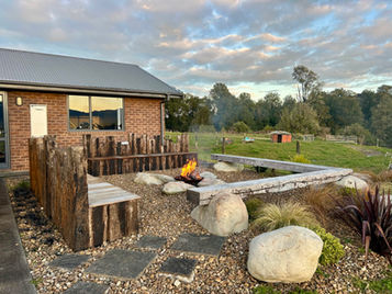 Outdoor fire pit area with stone-carved, aged hardwood benches, kwila seating on railway sleepers, surrounding planting, and a central fire pit built by Heartland Landscapes in Christchurch.