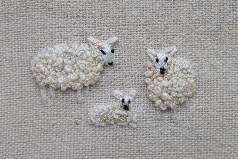 A family of 2 sheep and a lamb embroidered on a beige cotton background. Their fleece has been created with French knots.