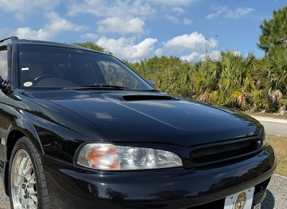 The front fascia of a 1996 Subaru Legacy Wagon GT-B, featuring its signature twin-turbo hood scoop, rectangular headlights, and a bold Subaru grille.